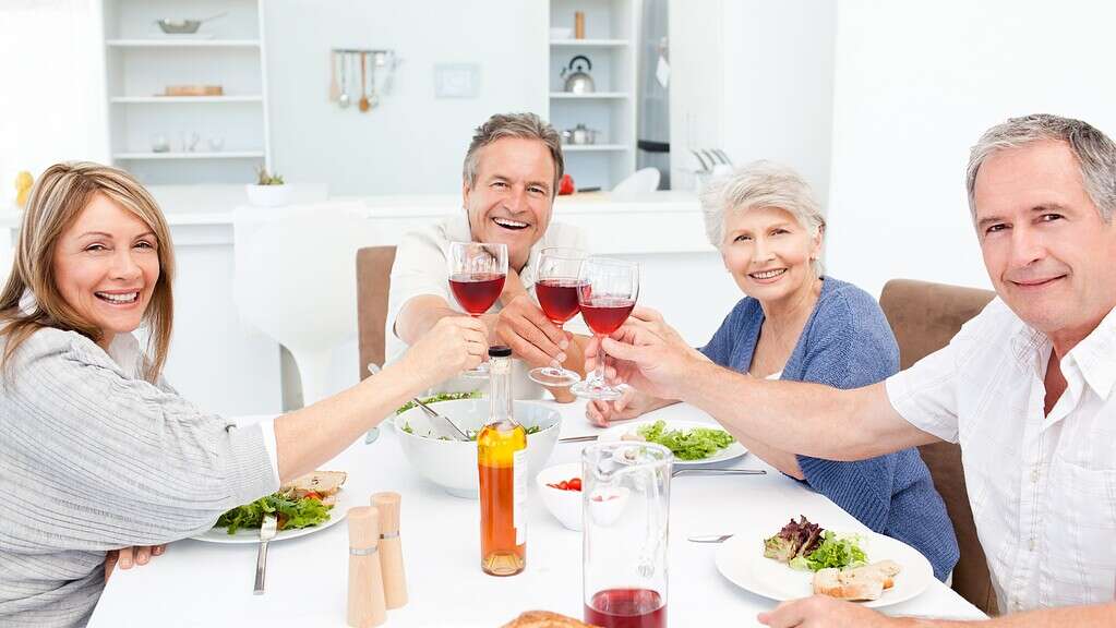 Personas mayores disfrutando de una comida en familia, brindando con vino tinto en un ambiente acogedor y moderno, promoviendo el bienestar y la convivencia en la tercera edad.